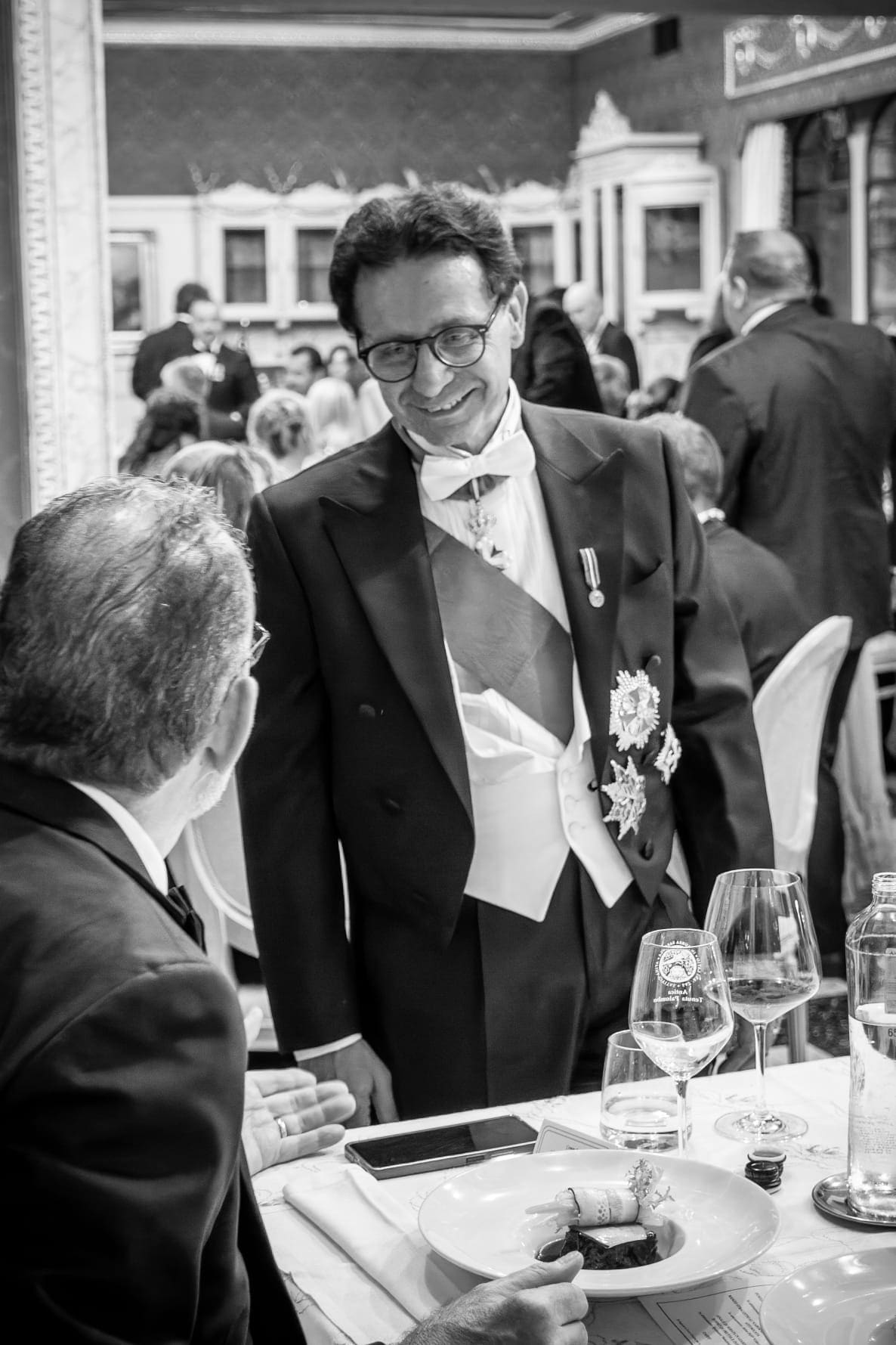 A member in white tie greeting a guest at a dinner table, in black and white