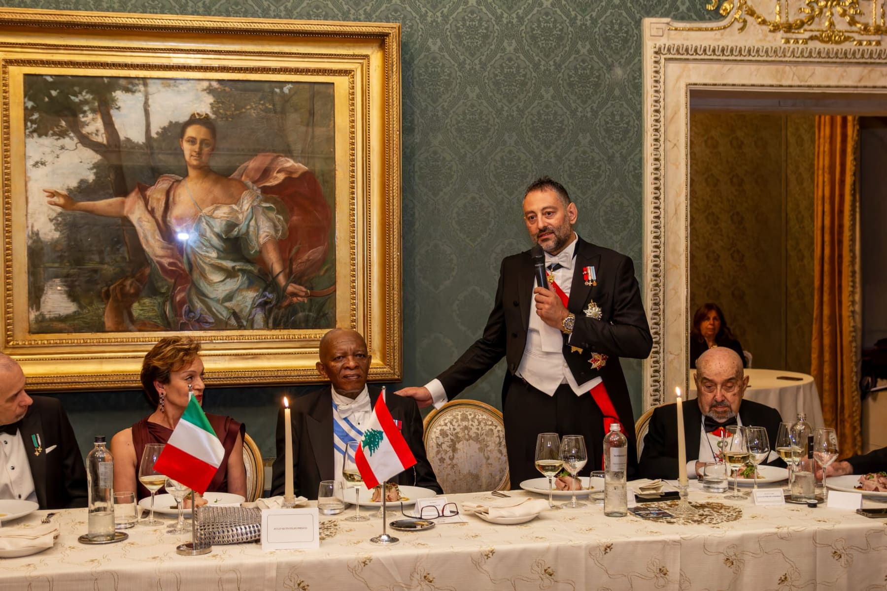A member rising to speak at a candlelit dinner beneath a painting
